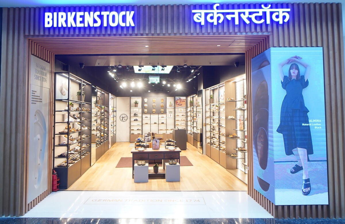 Birkenstock shoe store storefront with illuminated BIRKENSTOCK signage in English and Hindi, showing an open interior with shelving, display tables, and a large vertical digital advertisement panel on the right.